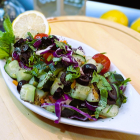 Abu Mubarak Authentic Arabic Salad in a white oval dish, featuring chopped cucumbers, cherry tomatoes, black olives, and red cabbage, garnished with fresh coriander and a lemon wedge.
