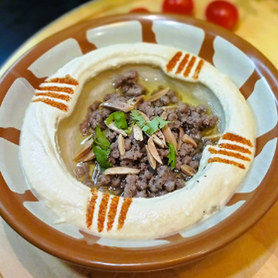 Abu Mubarak Authentic Beef Hummus in a traditional clay bowl, featuring smooth hummus topped with sautéed minced beef, toasted almonds, fresh coriander, and a drizzle of olive oil.
